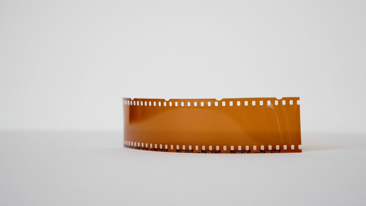 A curled strip of 35mm orange film on a white background, sharp sprocket holes — a stand-in for rendering video one frame at a time (photo by Markus Spiske, CC0, rawpixel)