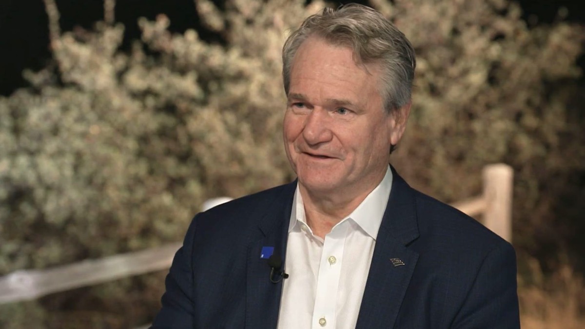 A man in a suit speaking in an interview in front of a wooden fence