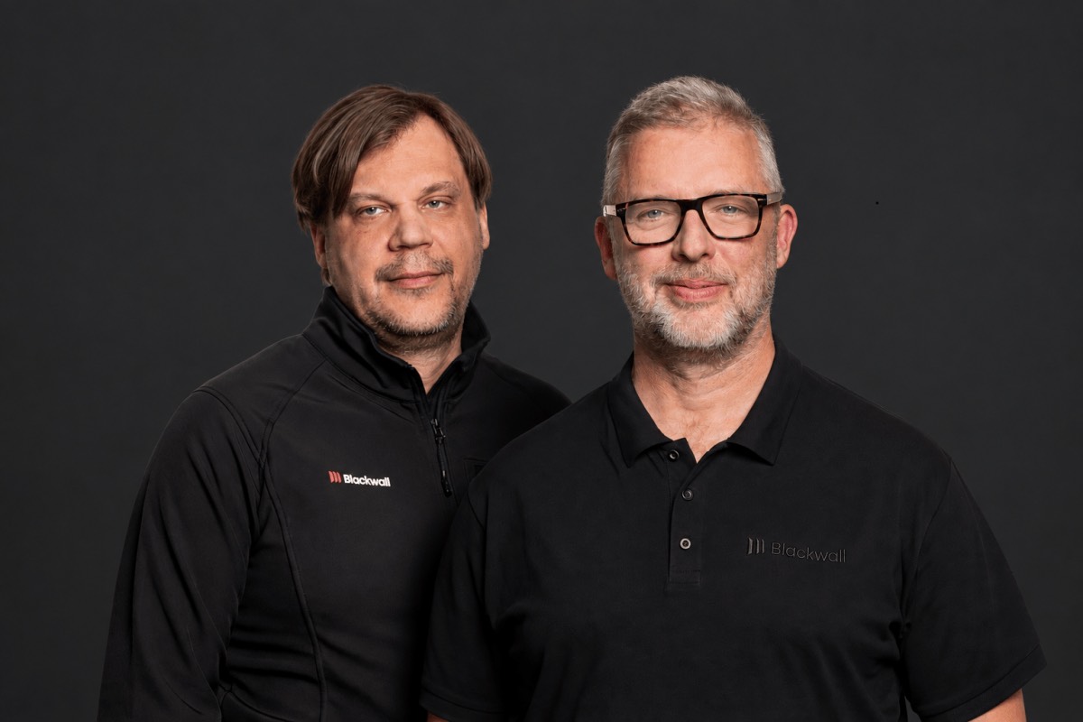 Two men posing against a dark backdrop, both in black tops with a small company logo
