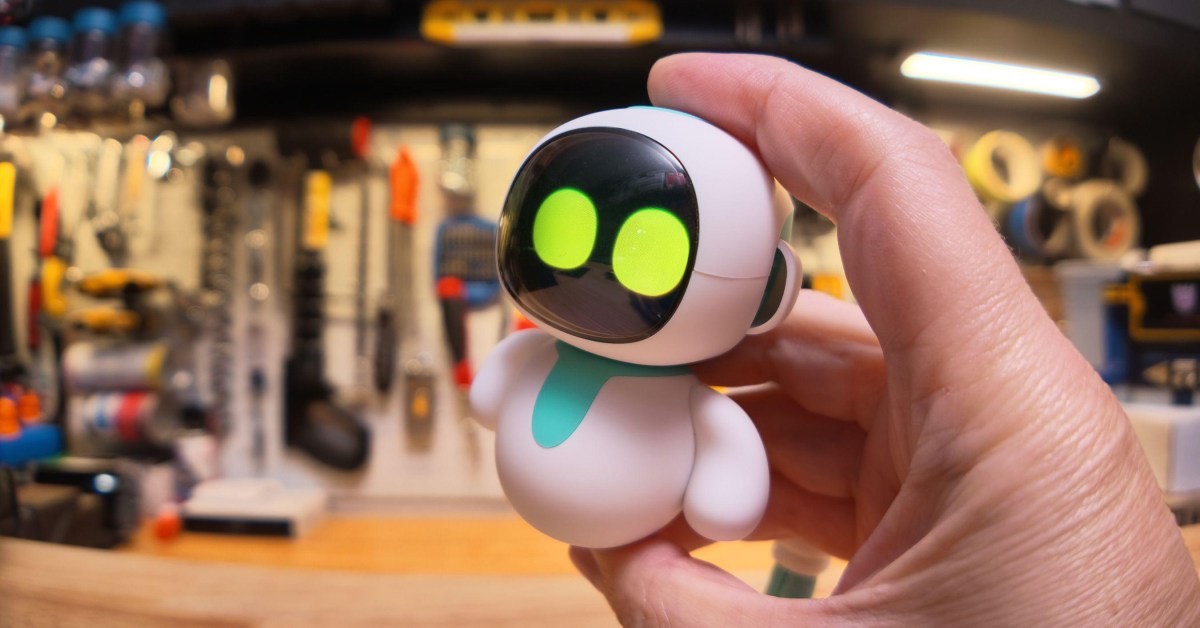 A hand holds the desktop Eiliko robot, its green eyes lit, in front of a workshop pegboard