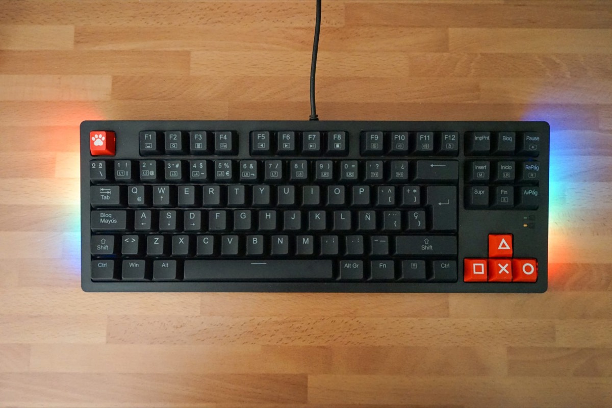 A black mechanical keyboard with RGB backlighting on a wooden desk, its arrow cluster swapped for red game-pattern keycaps