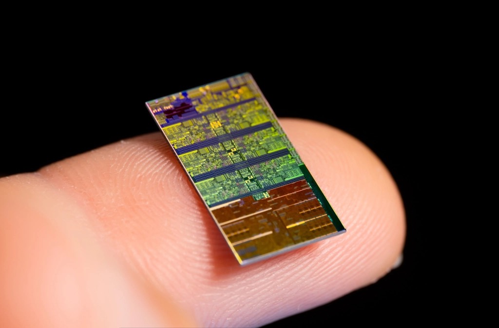 A bare silicon die resting on a fingertip, its circuit texture visible