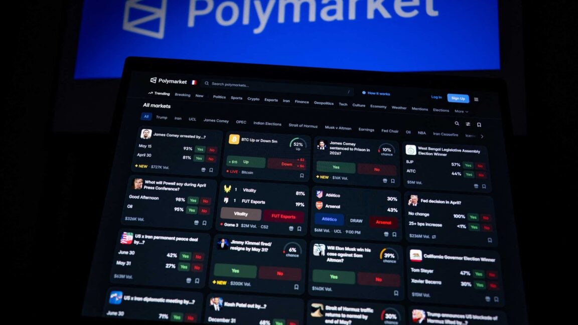 A Polymarket landing page projected on a wall above an open laptop showing a grid of prediction markets in a dark room