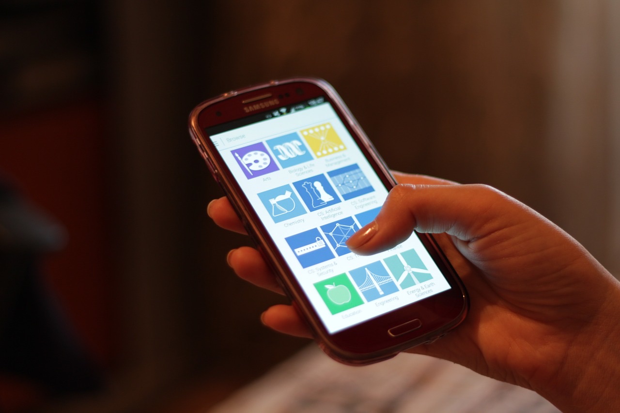 A hand holds an Android phone with a grid of app icons on screen, a second finger about to tap