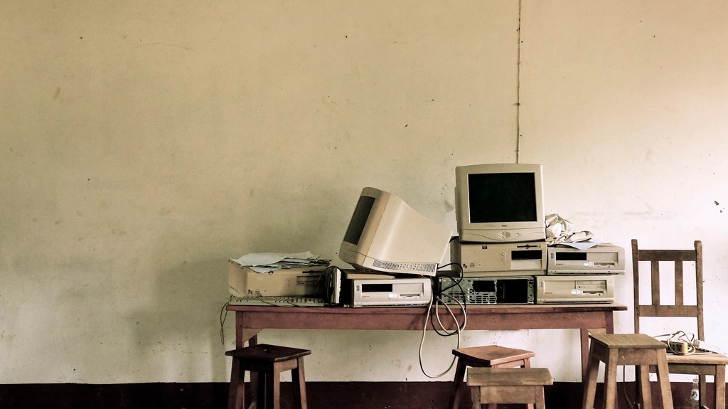 Old CRT monitors and legacy PC towers piled on a wooden table against a bare wall