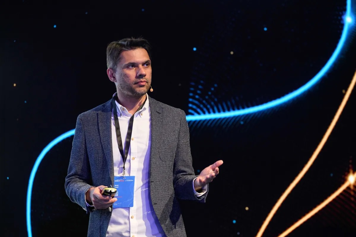 A man speaking on a conference stage against a dark backdrop with blue and orange light arcs