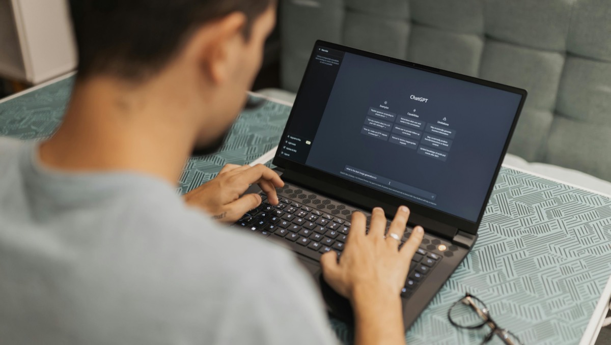 A laptop screen showing the ChatGPT interface while a user types on the keyboard, glasses resting on the desk beside it