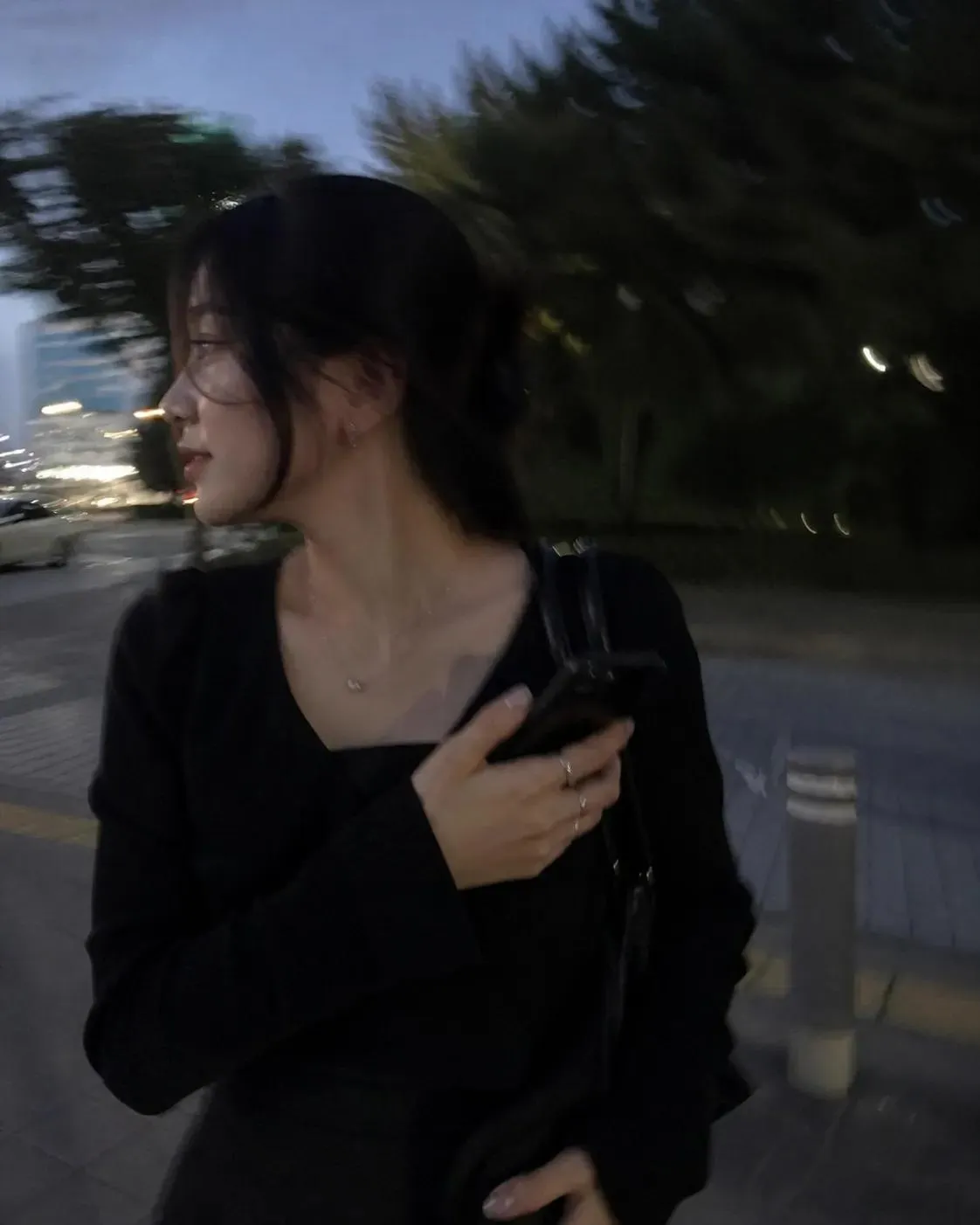 A candid night-street snapshot look: a young woman in black glances back over her shoulder, hair lifted by wind, the frame carrying motion blur and uneven streetlight